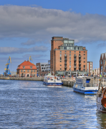 Eine Kogge liegt an der Hafenkante. Dahinter der Hafen von Wismar.