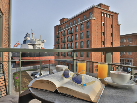 Buch und Orangesaftgläser auf einem Balkontisch mit Blick auf die Speichergebäuden in Wismar am Alten Hafen